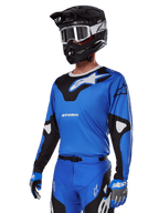 Ensemble d'équipement Alpinestars 2025 Racer Veil, maillot de motocross bleu avec graphismes noirs et blancs, porté avec un pantalon assorti, casque noir, lunettes blanches et gants bleus, tenue technique pour la course tout-terrain