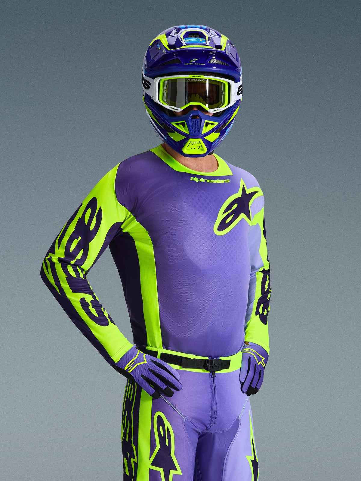 Pilote de motocross portant le maillot Alpinestars Racer Portl 2026, violet et jaune fluo, casque assorti violet et jaune avec lunettes blanches, gants violets et jaunes, tenue complète coordonnée pour la course tout-terrain