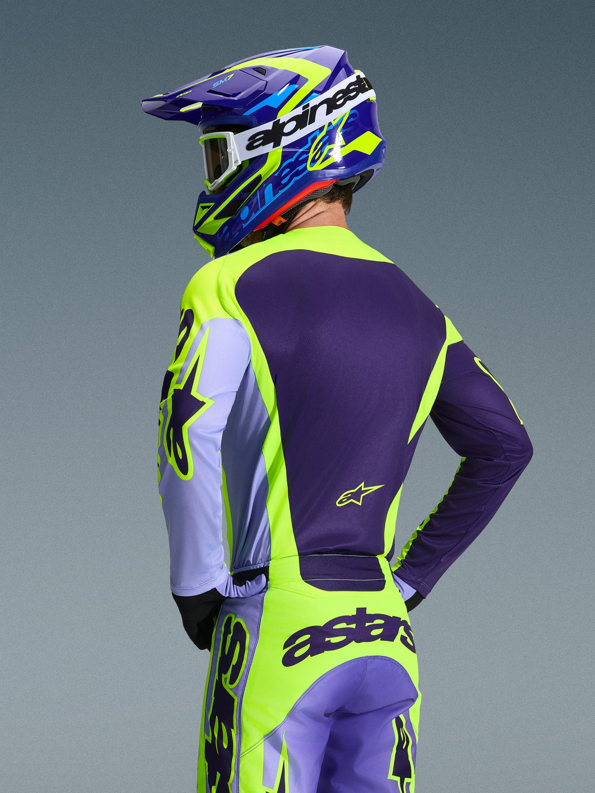 Pilote de motocross vu de dos portant le maillot Alpinestars 2026 Racer Portl et le pantalon assorti, coloris violet et jaune fluo avec accents lavande, casque bleu et jaune coordonné, équipement de course conçu pour la performance tout-terrain