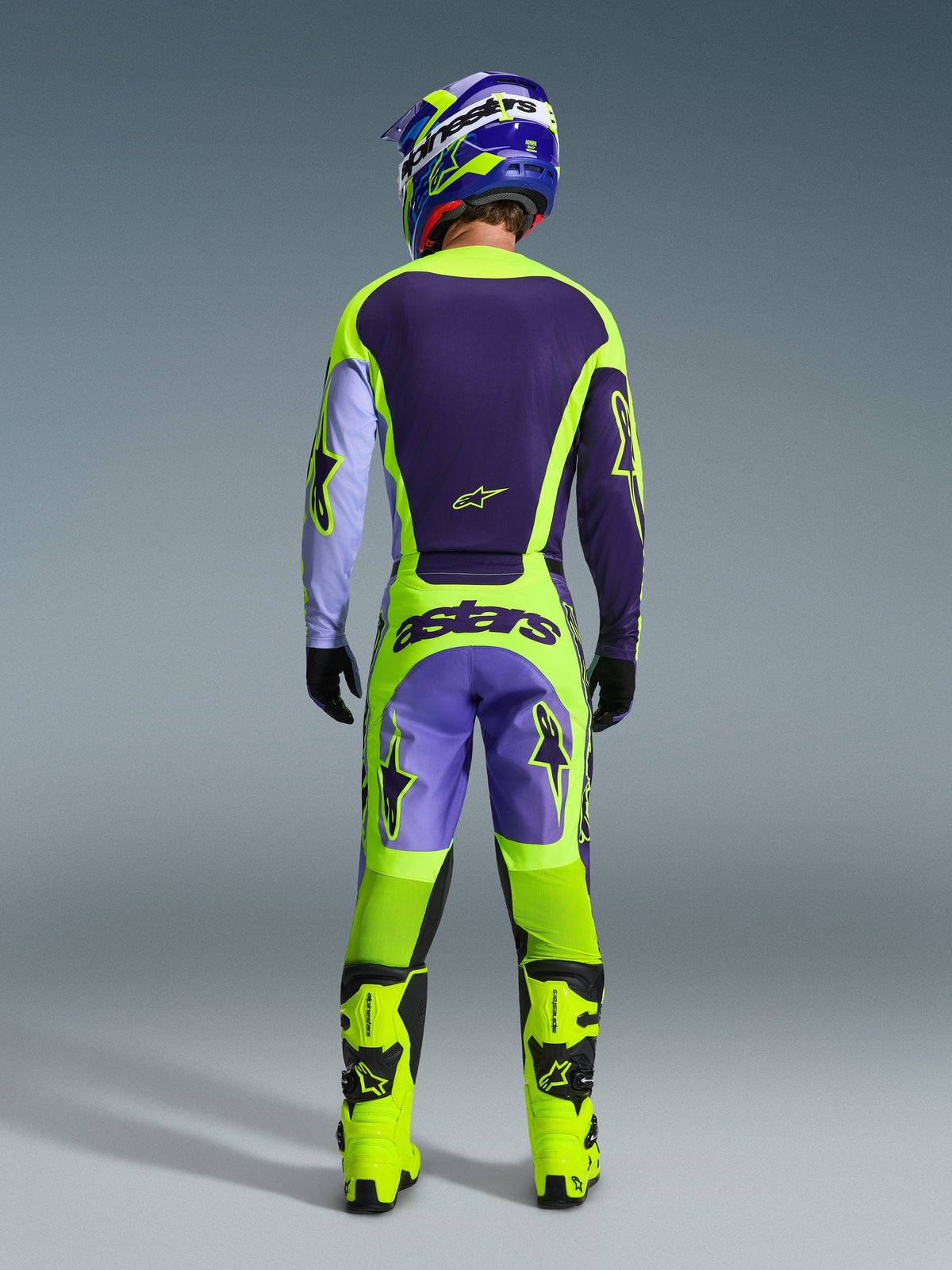 Pilote de motocross vu de dos portant le maillot Alpinestars Racer Portl 2026 en violet et jaune fluo, associé à un pantalon de motocross assorti, des bottes jaune fluo et un casque bleu et jaune, présentant une tenue complète tout-terrain.