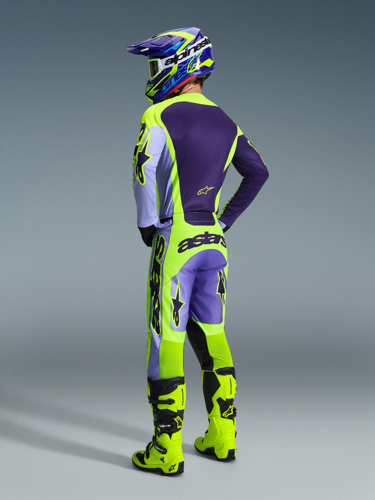 Pilote de motocross portant le maillot et le pantalon Alpinestars Racer Portl 2026, ensemble d'équipement violet et jaune fluo, vue de dos avec casque assorti et bottes jaune néon, tenue de course tout-terrain professionnelle coordonnée.