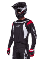 Pilote de motocross portant l'ensemble d'équipement Alpinestars Fluid Haul 2025, maillot noir avec empiècements rouges aux épaules et graphismes blancs, pantalon noir assorti avec logos étoiles blancs, gants rouges et noirs, casque noir avec détails blancs et lunettes, tenue de motocross coordonnée.