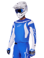 Pilote de motocross portant l'ensemble d'équipement Alpinestars Fluid Haul 2025 bleu et blanc, comprenant un maillot bleu avec détails blancs, un pantalon bleu assorti avec logos étoiles, un casque blanc avec lunettes bleues et des gants bleus et gris coordonnés, tenue de course tout-terrain complète