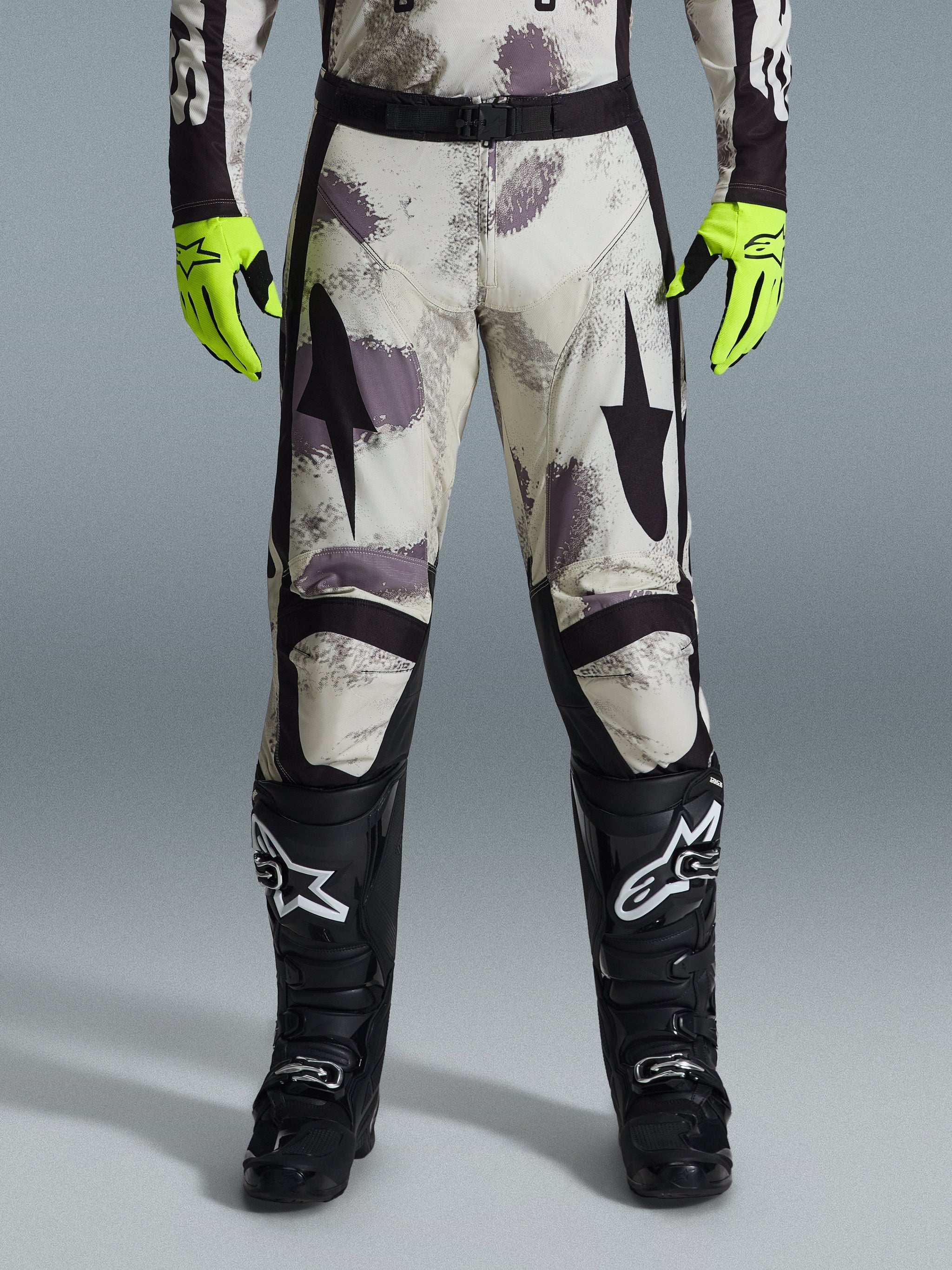 Alpinestars 2026 Racer Lahnd Pants, Motocross Pants, Sand Camo pattern with gray and purple abstract graphics, worn with neon yellow gloves and black boots, featuring black star logos on the thighs and a black waist belt, durable off-road racing gear