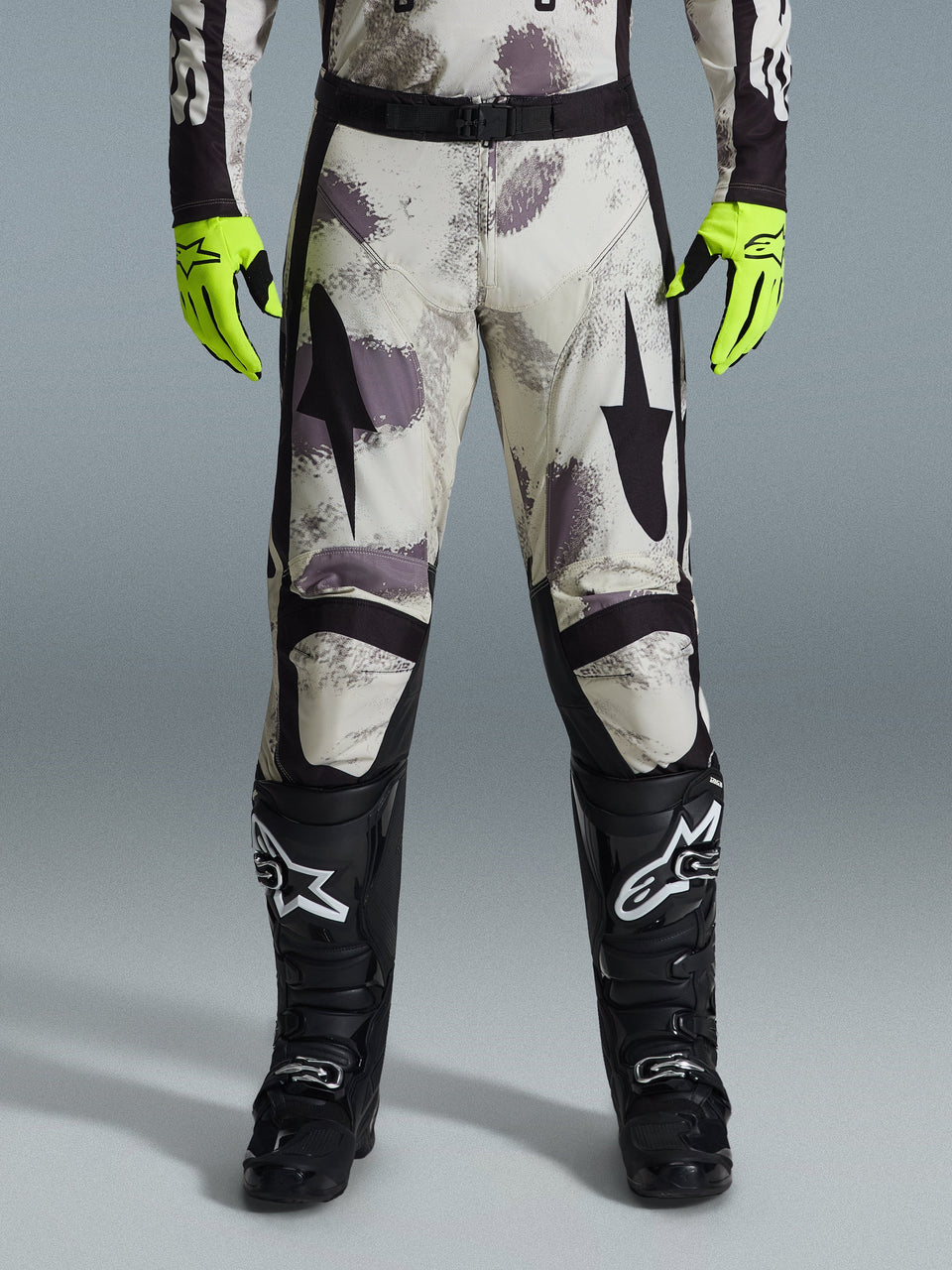 Pantalon Alpinestars Racer Lahnd 2026, pantalon de motocross, motif camouflage sable avec graphismes abstraits gris et violets, porté avec des gants jaune fluo et des bottes noires, avec des logos étoiles noirs sur les cuisses et une ceinture noire à la taille, équipement de course tout-terrain durable