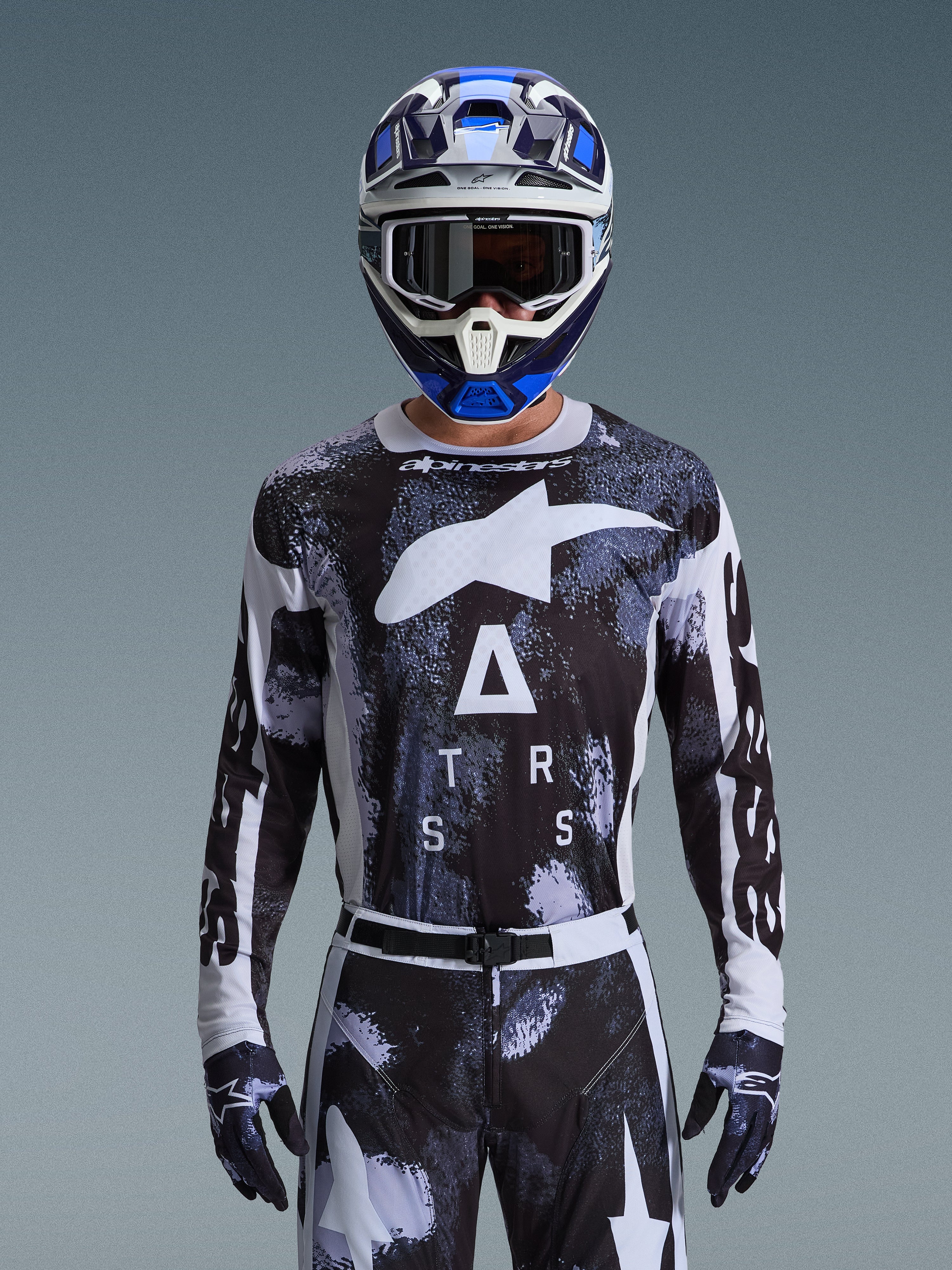 Pilote de motocross portant l'ensemble d'équipement Alpinestars Racer Lahnd 2026, maillot au motif camouflage abstrait noir et blanc avec grands logos blancs et lettrage « ATRS », pantalon et gants assortis, casque bleu et blanc avec masque, tenue de course haute performance pour le motocross tout-terrain