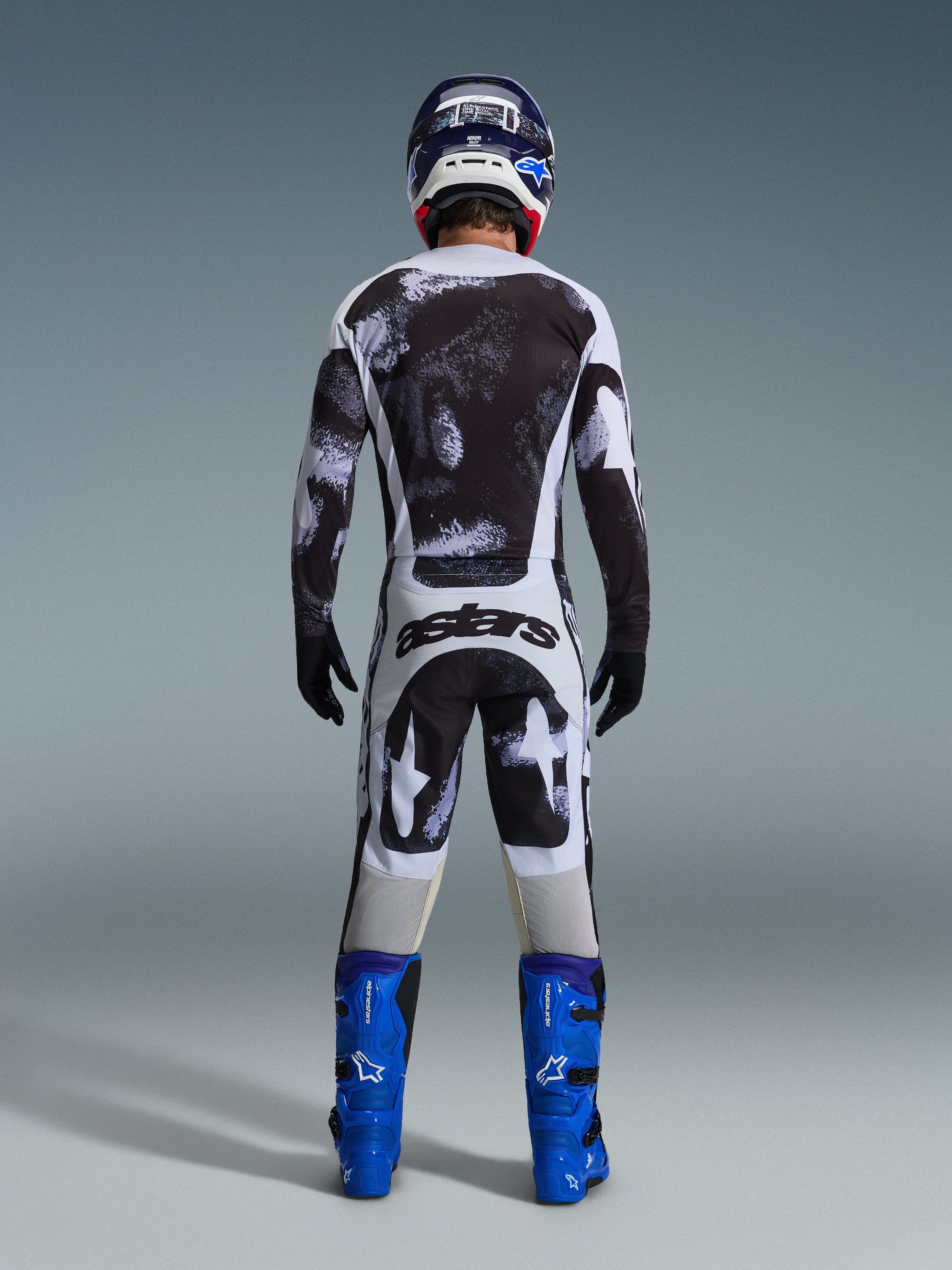 Pilote de motocross vu de dos portant l'ensemble Alpinestars 2026 Racer Lahnd, maillot et pantalon aux motifs abstraits noirs et blancs, marquage « astars » sur l'assise, avec bottes bleues et casque assorti, équipement professionnel de course tout-terrain.