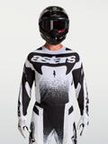 Maillot Alpinestars Techstar Swan, maillot de motocross pour homme, blanc et noir, coupe athlétique en tissu extensible léger avec perforations laser, porté par un mannequin avec casque et gants