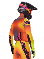 Pilote de motocross vu de trois-quarts arrière portant l'ensemble d'équipement Alpinestars Supertech Lipan 2025, présentant un dégradé jaune, orange et rouge avec des logos noirs et roses, casque noir avec accents roses et lunettes orange, tenue de course tout-terrain professionnelle.