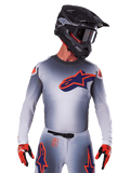 Maillot Alpinestars Supertech Lipan 2025, maillot de motocross, gris avec des accents orange et bleu marine, haut de performance à manches longues avec détails de ventilation perforés, porté avec un casque noir et des gants rouges, design de course épuré