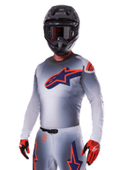 Pilote de motocross portant l'ensemble Alpinestars Supertech Lipan 2025, maillot et pantalon gris avec accents bleu marine et orange, casque noir mat avec lunettes, gants rouges et noirs, équipement de performance athlétique pour la course tout-terrain