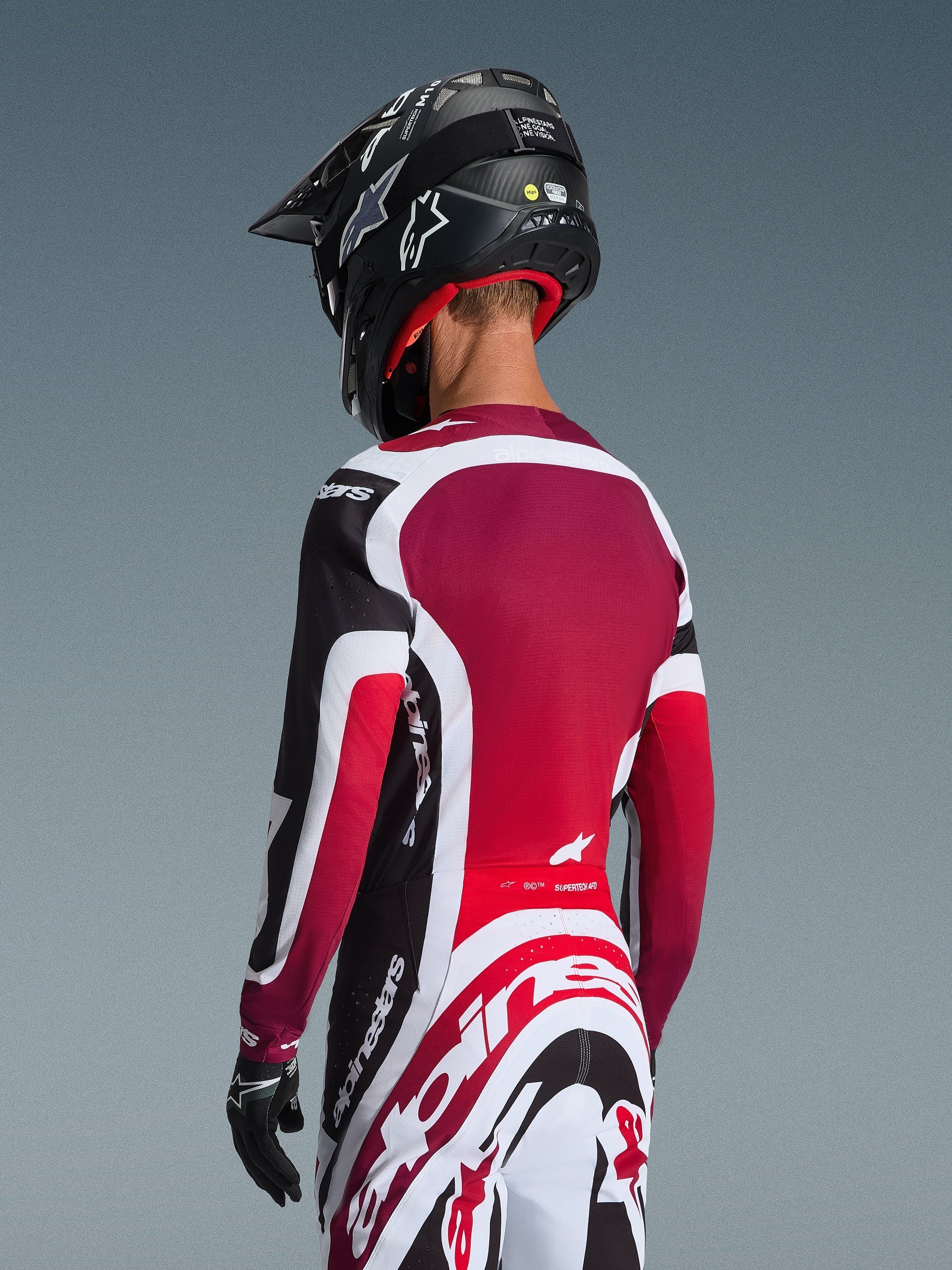 Vue arrière d'un pilote de motocross portant le maillot Supertech AFD Vista 2026 noir, rouge et blanc, coordonné avec un pantalon assorti et un casque noir, arborant le logo Alpinestars, des panneaux en mesh rouge et une coupe racing athlétique.