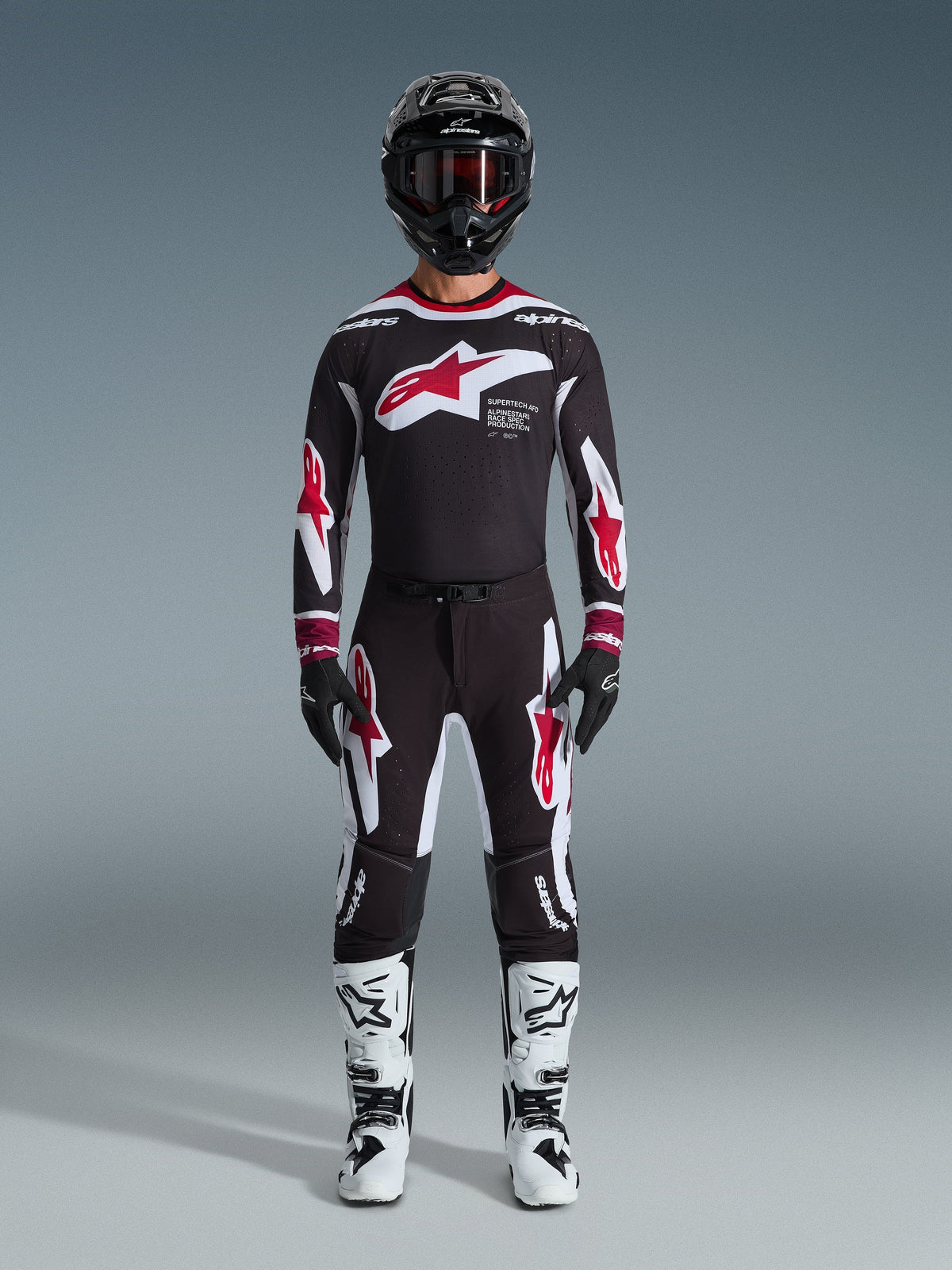 Motocross rider wearing Alpinestars 2026 Supertech AFD Vista Jersey, Black Red and White racing gear, matching black pants with red star logos, white and black boots, black helmet with goggles, professional off-road racing kit.