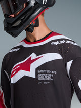 Alpinestars Supertech AFD Vista Jersey 2026, gros plan d'un pilote de motocross portant un maillot noir avec des accents rouges et blancs, arborant un grand logo Alpinestars blanc et rouge, des perforations de ventilation découpées au laser et une coupe racing professionnelle, assorti à un casque noir.