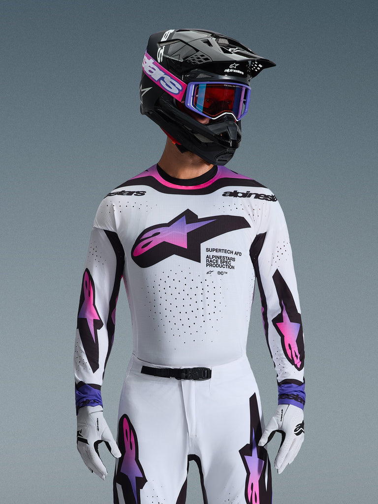 Pilote de motocross portant le maillot Supertech AFD Vista 2026 en blanc, violet et noir, comprenant un casque noir avec marquage rose, des lunettes violettes et des gants blancs assortis, équipement à coupe athlétique conçu pour la performance en compétition tout-terrain professionnelle