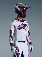 Motocross rider wearing Alpinestars 2026 Supertech AFD Vista Gear Set, white jersey and pants with pink and purple gradient star logos, black helmet with purple accents, purple goggles with red-tinted lenses, and matching white gloves, coordinated professional off-road racing outfit.