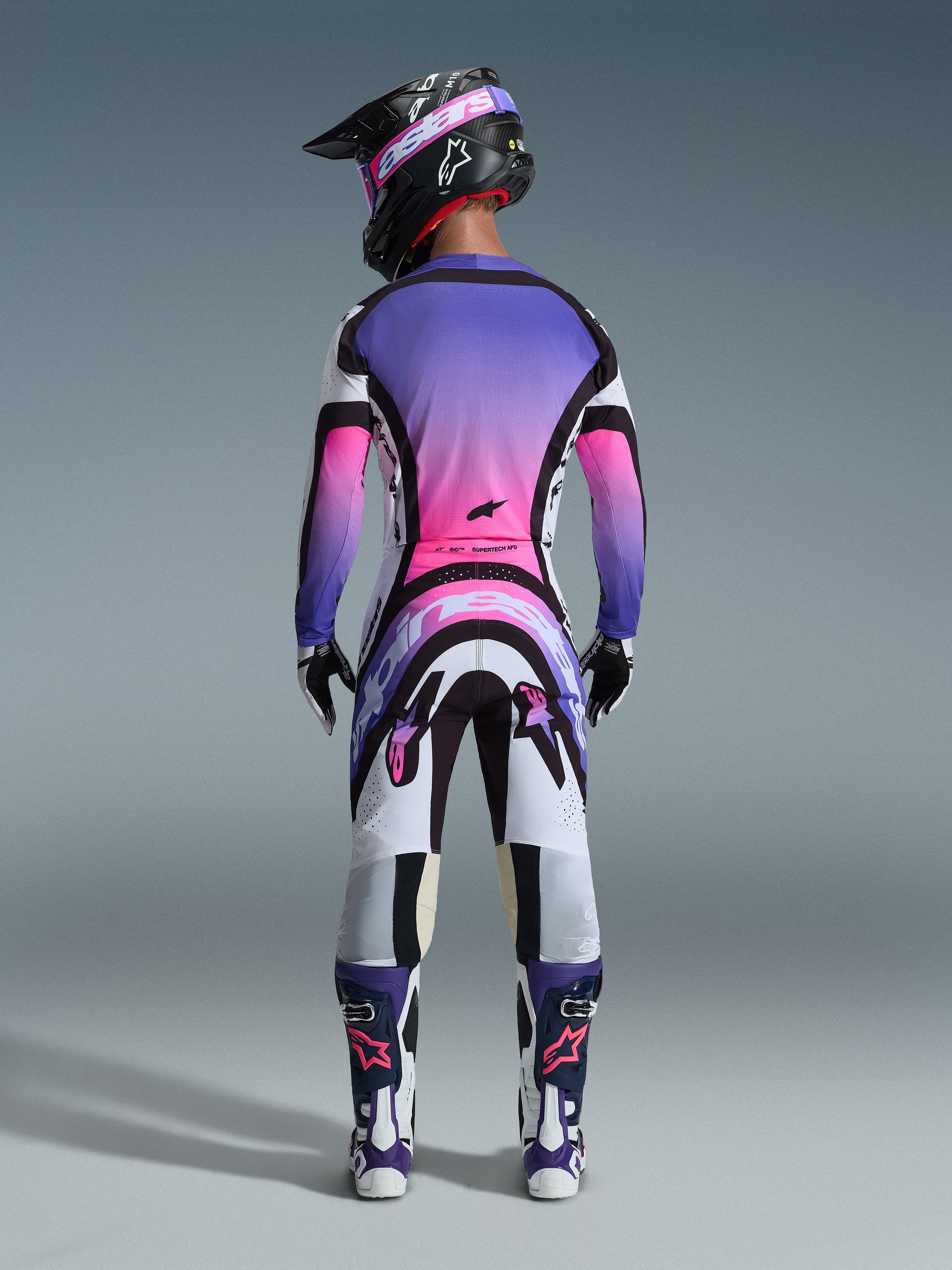 Rear view of a motocross rider wearing the Alpinestars 2026 Supertech AFD Vista Gear Set, jersey and pants featuring a vibrant white, purple, and pink gradient with black accents, black helmet with purple goggles, matching purple and white boots, coordinated professional racing outfit for off-road performance.