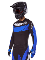 Pilote de motocross portant la tenue Alpinestars Techstar Dreem 2025, maillot noir avec empiècements bleus au style perforé et logos blancs, pantalon noir et bleu assorti, casque de course noir et gants bleus, tenue de performance coordonnée pour le motocross tout-terrain.