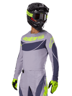 Pilote de motocross portant le maillot Alpinestars Techstar Dreem 2025, gris et gris foncé avec logos vert citron, pantalon assorti, casque noir avec accents vert citron, gants vert citron et noirs, tenue coordonnée pour la course tout-terrain