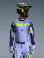 Vue de face d'un pilote de motocross portant l'ensemble d'équipement Supertech Scenz 2026, maillot violet clair et bleu foncé avec marquage 'astars' jaune fluo éclatant, pantalon et gants assortis avec détails fluo, casque noir et lunettes jaune fluo, photo de studio sur fond gris.