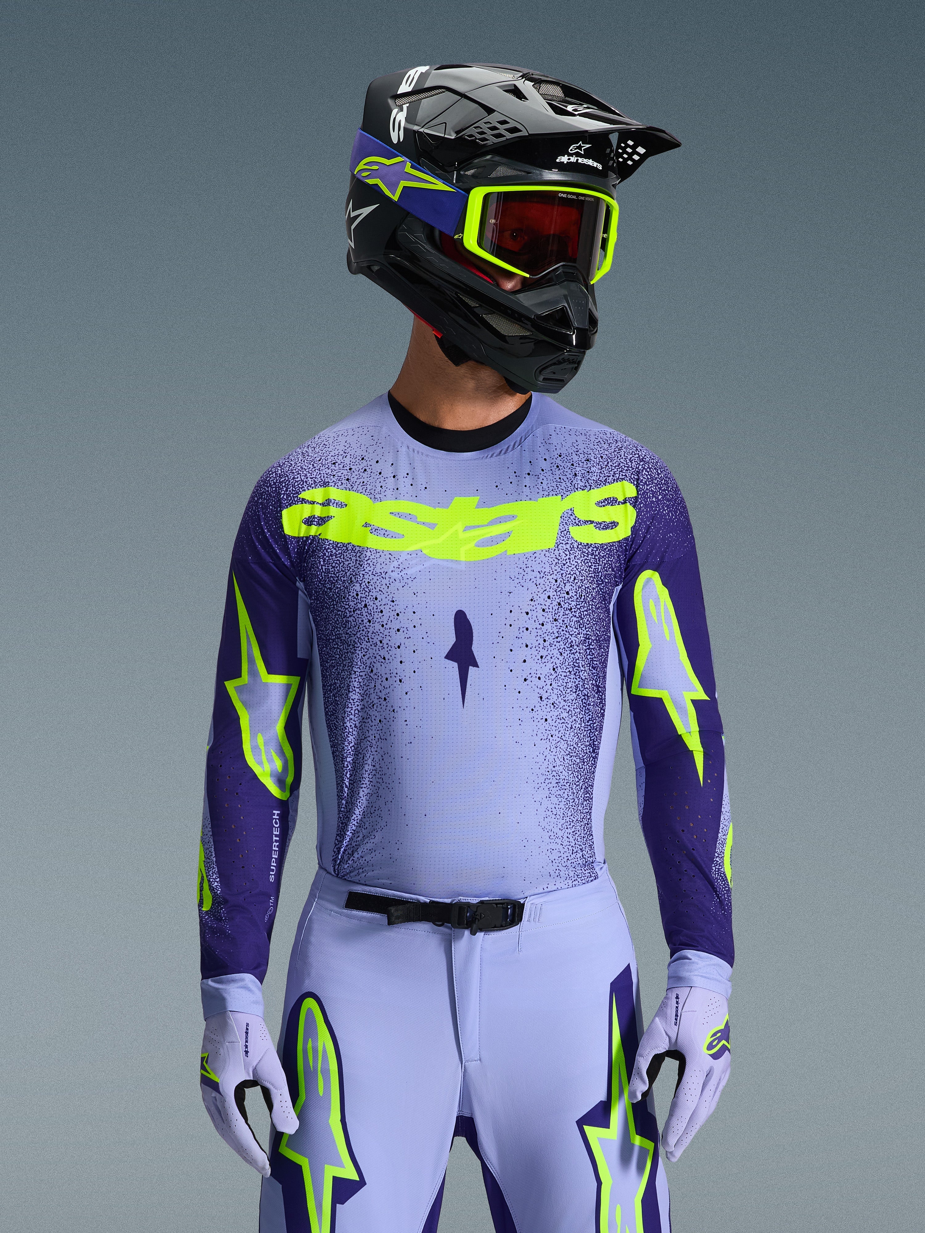 Vue de face d'un pilote de motocross portant l'ensemble d'équipement Supertech Scenz 2026, maillot violet clair et bleu foncé avec marquage 'astars' jaune fluo éclatant, pantalon et gants assortis avec détails fluo, casque noir et lunettes jaune fluo, photo de studio sur fond gris.