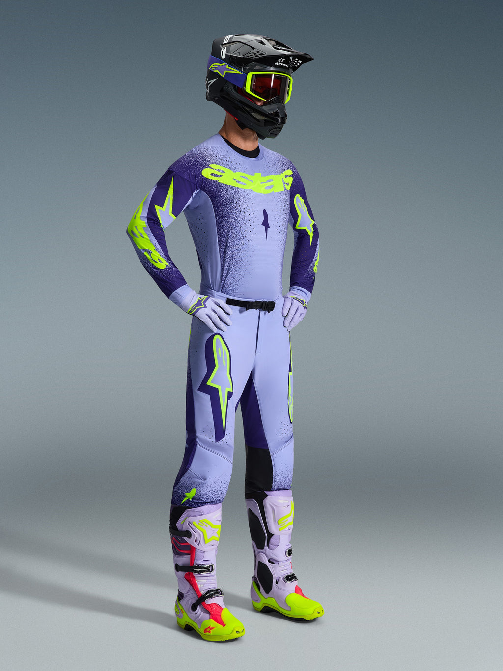 Ensemble d'équipement Alpinestars 2026 Supertech Scenz, pilote de motocross portant une tenue de course complète, maillot violet clair avec manches violet foncé et accents jaune fluo, pantalon lavande assorti, casque noir avec lunettes jaune fluo, et bottes lavande avec détails vert fluo et rouges, tenue tout-terrain professionnelle coordonnée.