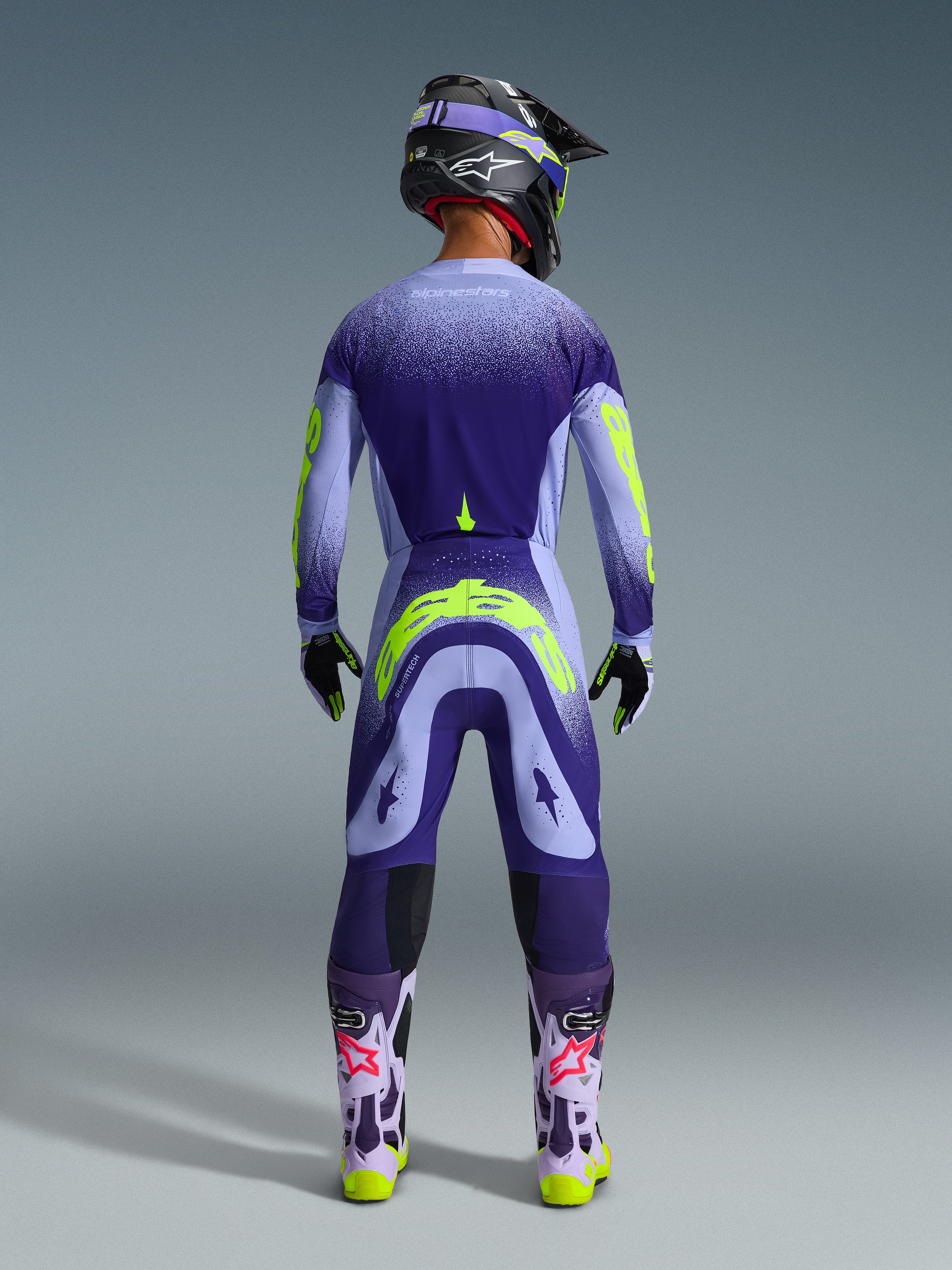 Alpinestars 2026 Supertech Scenz Gear Set, pilote de motocross vu de dos portant une tenue violette et lilas avec des accents jaune fluo, maillot et pantalon avec motif à points en dégradé, casque et lunettes assortis, bottes lilas clair avec logos roses et semelles jaune fluo, équipement de course professionnel