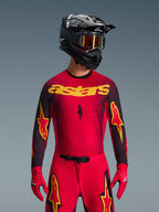 Alpinestars 2026 Supertech Scenz Gear Set, Motocross rider wearing a red jersey with yellow 'astars' branding and black speckled details, red pants with yellow star logos on the thighs, red racing gloves, and a black helmet with reflective yellow goggles, pro-level racing outfit for off-road performance