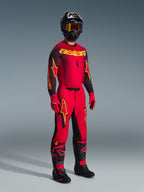 Motocross rider wearing complete racing gear, red jersey with black sleeves and yellow graphics, matching red pants with black panels and yellow logos, black helmet with yellow-lens goggles, black and red boots, red and black gloves, coordinated 2026 Supertech Scenz outfit for professional motocross racing
