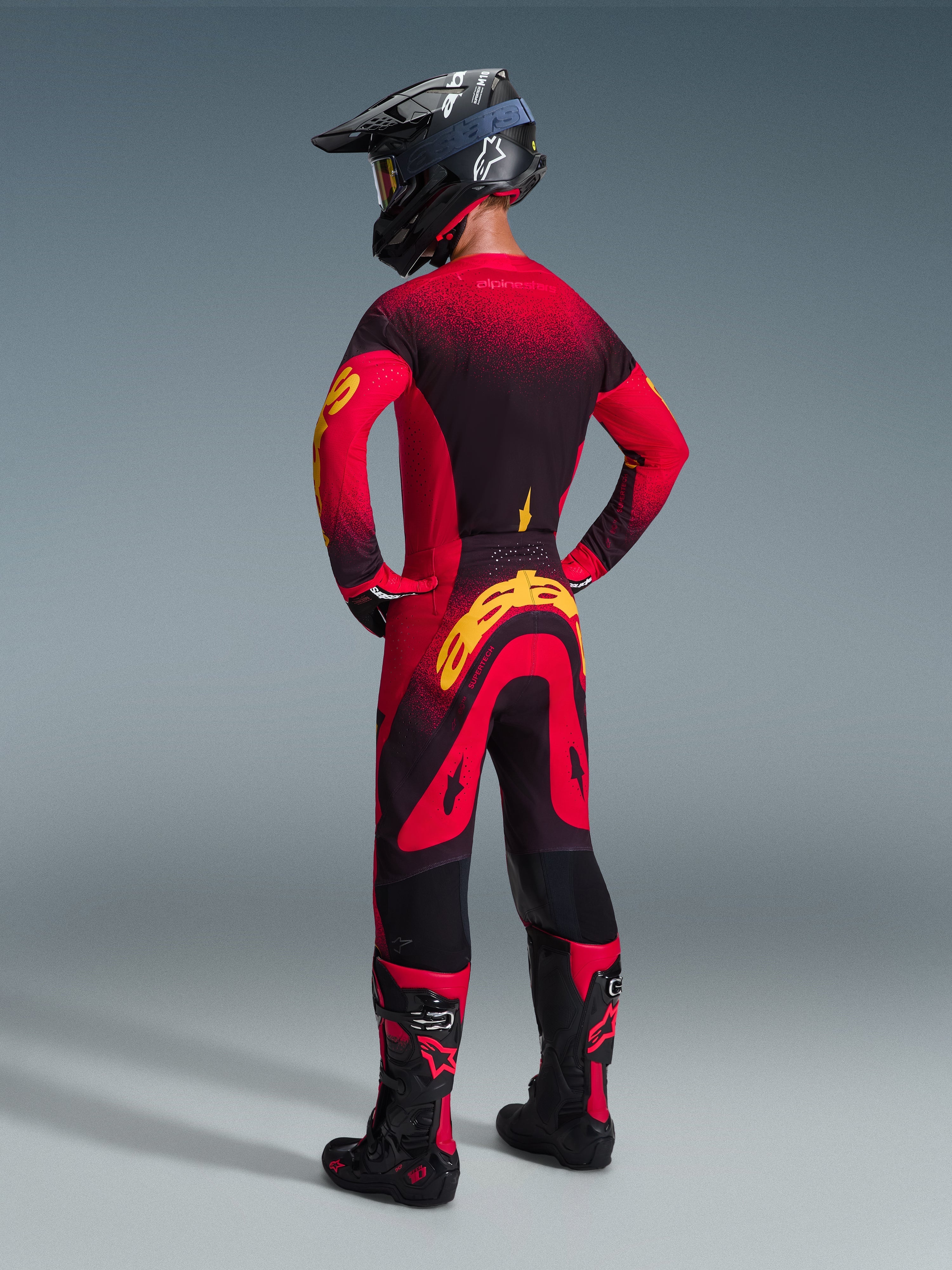Motocross rider wearing complete racing gear seen from the back, Alpinestars 2026 Supertech Scenz Jersey in Red Black and Yellow, matching motocross pants with yellow branding, black helmet, and black and red boots, coordinated outfit for professional off-road racing.