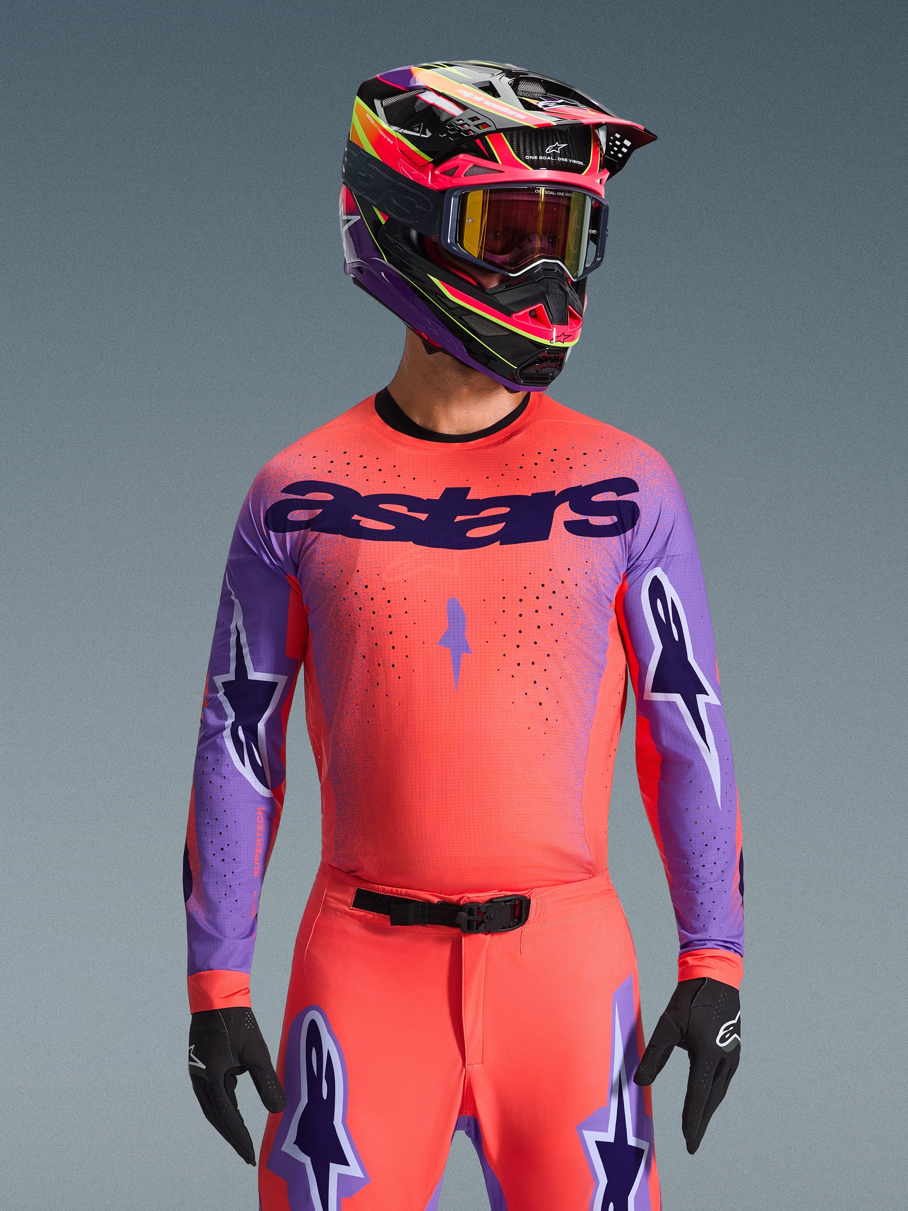 Maillot Alpinestars Supertech Scenz 2026, maillot de motocross, corail vif et violet, avec grand logo 'astars' et motifs d'étoiles sur les manches, porté avec un pantalon assorti et un casque multicolore, équipement tout-terrain haute performance