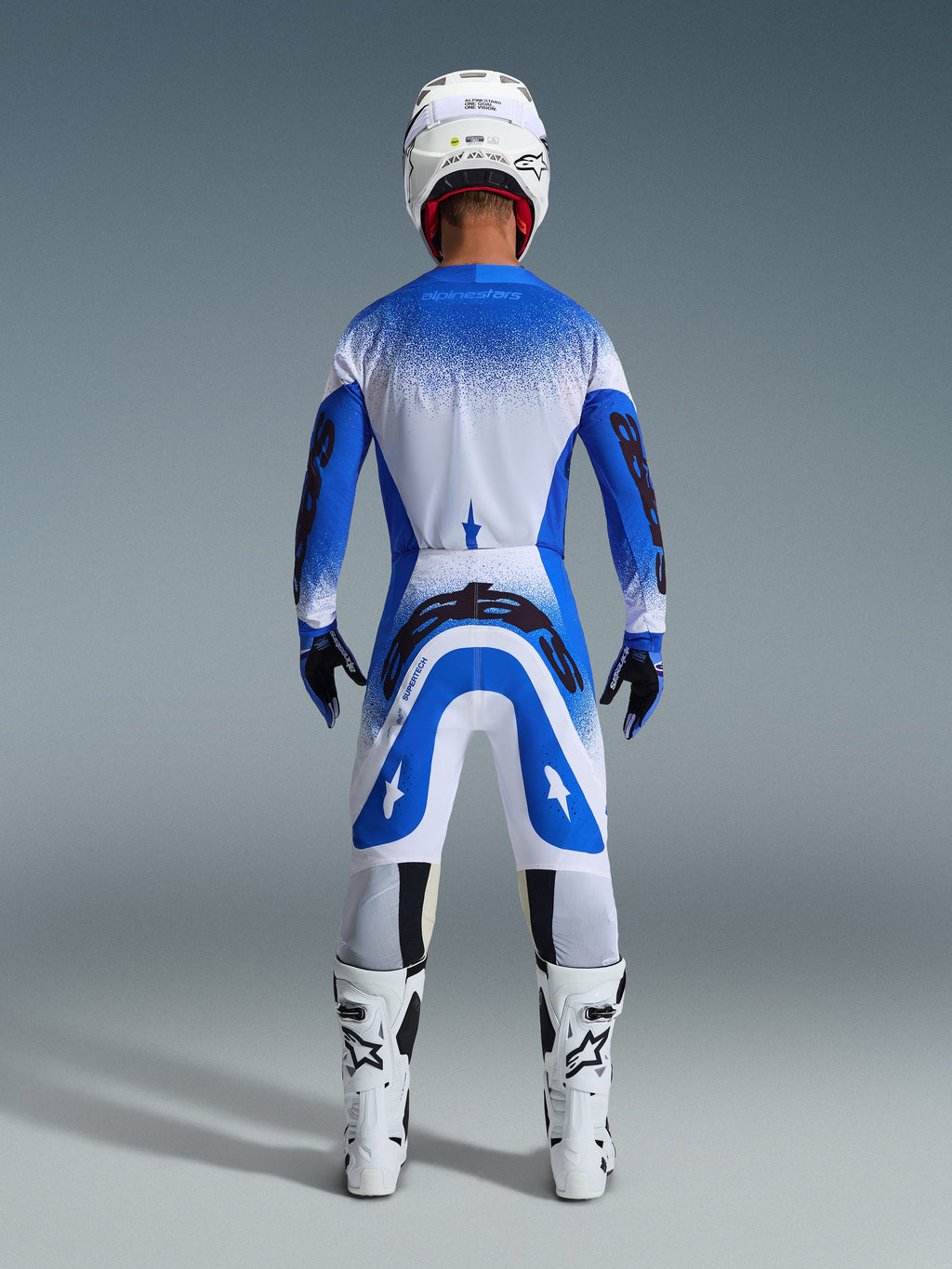 Motocross rider from the back wearing the Alpinestars 2026 Supertech Scenz Gear Set in Ucla Blue White, blue and white jersey with black branding, matching pants, white helmet, and white boots, coordinated pro-level racing gear for off-road motocross