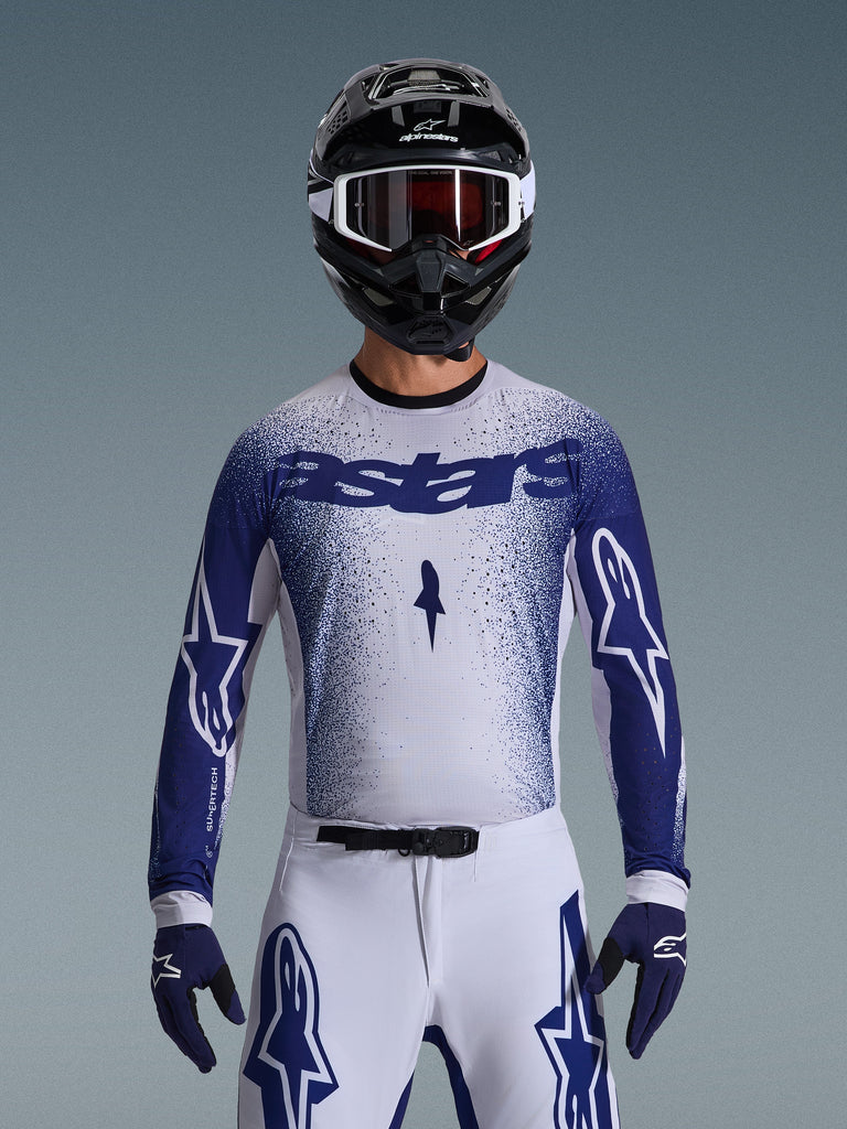 Pilote de motocross portant le maillot Alpinestars 2026 Supertech Scenz, gris clair et bleu foncé avec un motif dégradé moucheté et un marquage 'astars' audacieux. Porté avec un pantalon de motocross blanc assorti, des gants bleu foncé et un casque noir avec lunettes, tenue coordonnée pour la course tout-terrain professionnelle.