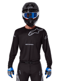 Maillot Alpinestars Racer Graphite 2025, équipement de motocross, noir avec logos gris, porté avec un casque noir assorti, des lunettes bleues et des gants bleus et gris, design de performance ajusté avec marquage sur la poitrine et les bras