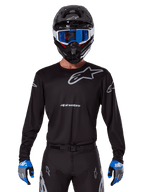 Maillot Alpinestars Racer Graphite 2025, équipement de motocross, noir avec logos gris, porté avec un casque noir assorti, des lunettes bleues et des gants bleus et gris, design de performance ajusté avec marquage sur la poitrine et les bras