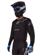 Pilote de motocross portant l'ensemble d'équipement Alpinestars Racer Graphite 2025, maillot noir avec logos Alpinestars gris, pantalon noir assorti avec accents gris, casque noir avec lunettes bleues et blanches, gants bleus et gris, tenue technique épurée pour la course tout-terrain