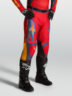 Alpinestars Pantalon Racer Hunt, pantalon de motocross pour homme rouge avec accents gris et corail, porté avec des bottes noires.