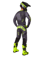 Pilote de motocross de dos portant le maillot et le pantalon Alpinestars Racer Hollow 2025 en coloris Dark Camo Yellow Fluo, avec un motif camouflage gris foncé et des accents jaune fluo, assorti à un casque noir et jaune et des bottes jaune fluo éclatantes.