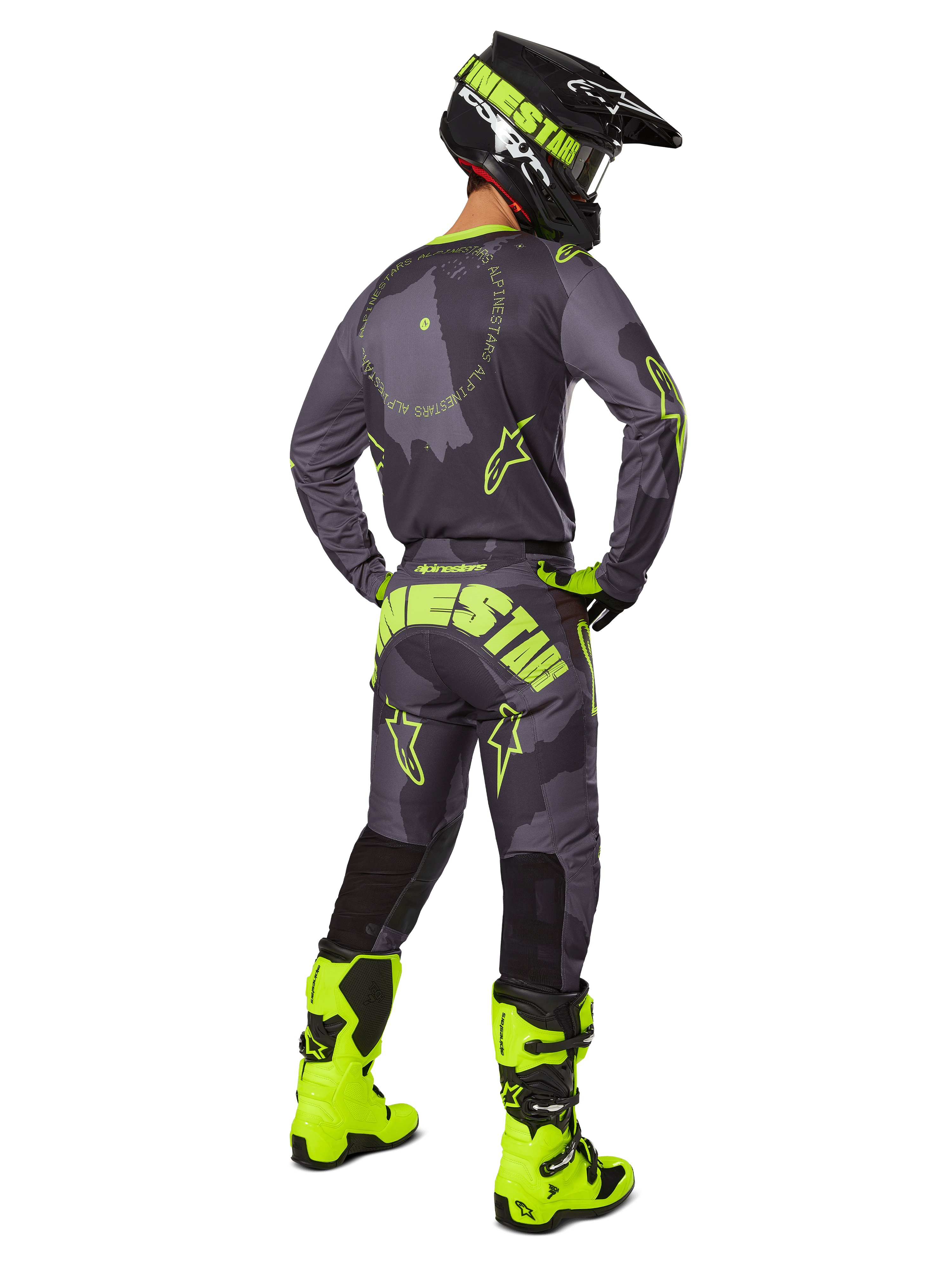 Pilote de motocross de dos portant le maillot et le pantalon Alpinestars Racer Hollow 2025 en coloris Dark Camo Yellow Fluo, avec un motif camouflage gris foncé et des accents jaune fluo, assorti à un casque noir et jaune et des bottes jaune fluo éclatantes.
