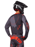 Maillot Alpinestars Racer Hollow 2025, vue arrière d'un pilote de motocross en tenue Gris Camo et Orange Fluo, avec un pantalon camouflage assorti arborant un grand lettrage Alpinestars orange et un casque noir, design d'équipement tout-terrain professionnel