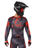 Alpinestars 2025 Racer Hollow Jersey, maillot de motocross, camouflage gris et orange fluo, porté avec une tenue assortie incluant un pantalon et des gants, doté d'un casque noir avec des graphismes blancs et des lunettes de protection, logo ASTARS audacieux, vêtement de sport de protection pour la course tout-terrain