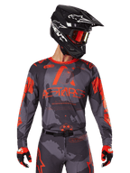 Alpinestars 2025 Racer Hollow Jersey, maillot de motocross, camouflage gris et orange fluo, porté avec une tenue assortie incluant un pantalon et des gants, doté d'un casque noir avec des graphismes blancs et des lunettes de protection, logo ASTARS audacieux, vêtement de sport de protection pour la course tout-terrain