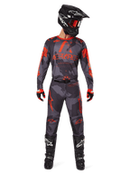 Pilote de motocross portant le maillot Alpinestars Racer Hollow 2025 et le pantalon assorti en Gris Camo Orange Fluo, tenue complète avec casque, lunettes, gants et bottes noirs, avec logos orange vifs et motif camouflage.