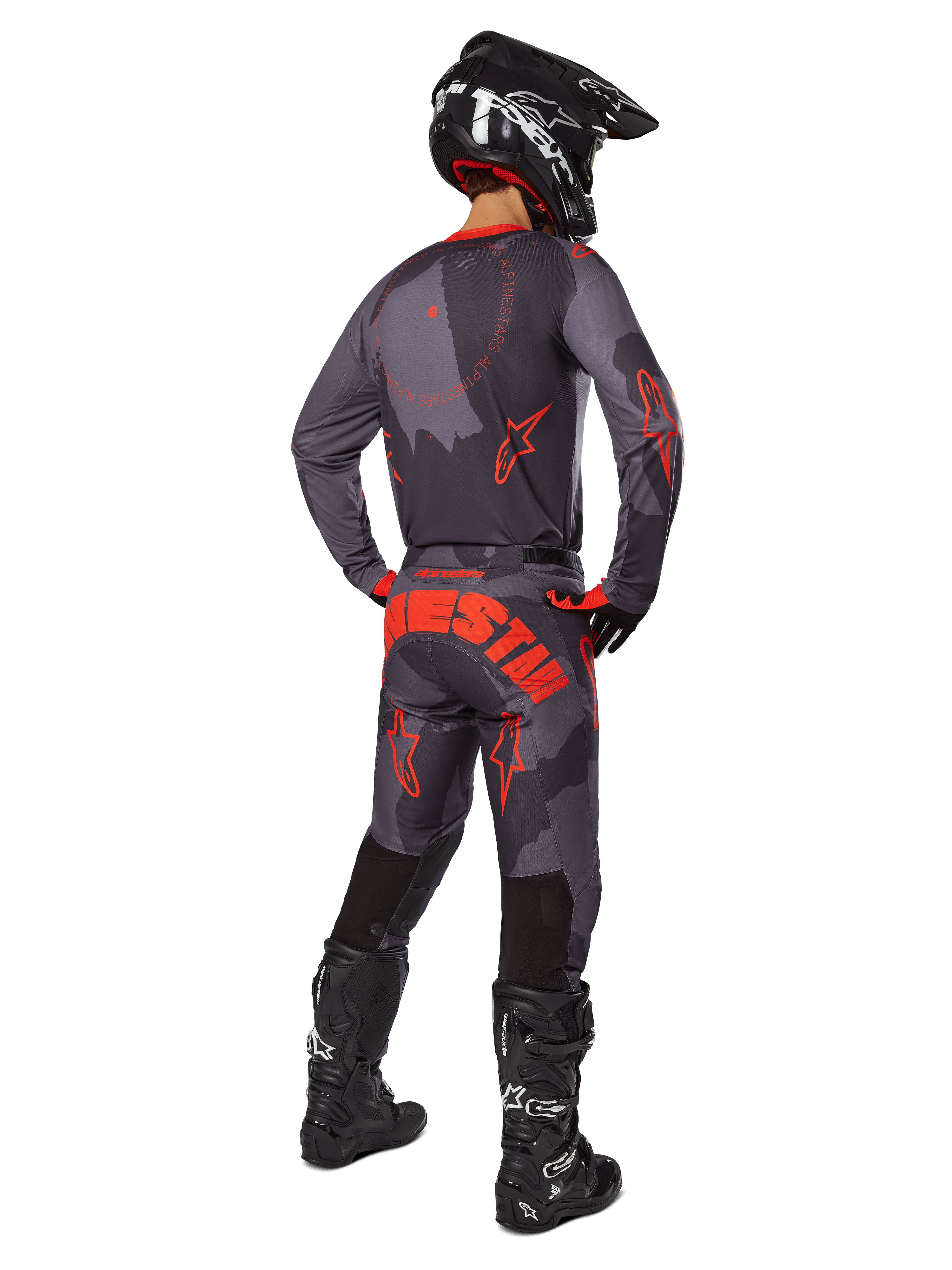 Pilote de motocross vu de dos portant le maillot Alpinestars 2025 Racer Hollow en Gris Camo Orange Fluo, pantalon camouflage assorti avec texte Alpinestars orange, casque noir et bottes noires, tenue coordonnée pour le tout-terrain