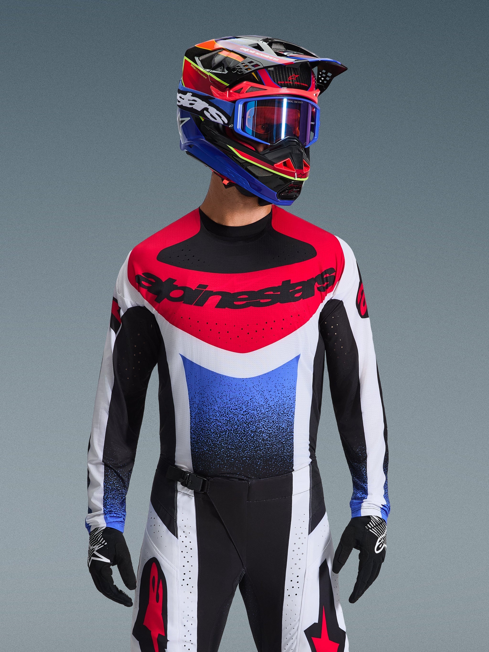 Alpinestars 2026 Techstar Knif Jersey, Motocross Jersey, Red White and Blue, featuring a bold chest logo and gradient torso, worn by a rider with a matching multi-colored helmet, blue goggles, and black gloves, professional off-road racing gear.