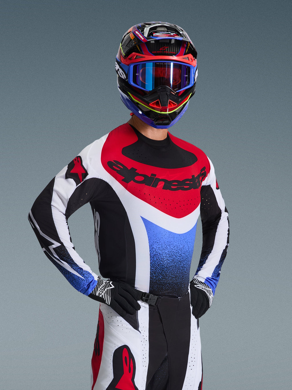 Alpinestars 2026 Techstar Knif Gear Set, pilote de motocross en maillot de course rouge, blanc et bleu avec accents noirs, casque et lunettes multicolores assortis, gants noirs, équipement professionnel pour la compétition de motocross.