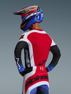 Pilote de motocross de dos portant la tenue Alpinestars Techstar Knif 2026, maillot rouge avec empiècements latéraux blancs et manches noires aux poignets mouchetés de bleu, pantalon rouge et blanc avec logo Alpinestars noir sur l'assise, casque bleu et rouge coordonné avec lunettes.