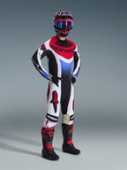 Pilote de motocross portant l'ensemble d'équipement Alpinestars 2026 Techstar Knif, maillot composé de panneaux noirs, blancs et rouges avec un dégradé bleu sur la poitrine, pantalon blanc avec logos Alpinestars rouges et accents noirs, bottes noires et rouges, tenue de course tout-terrain professionnelle complète.