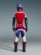 Pilote de motocross portant la tenue complète Alpinestars Techstar Knif 2026, vue de dos, comprenant un maillot rouge, blanc et noir avec des accents dégradés bleus sur les manches, un pantalon blanc et noir assorti avec un marquage rouge audacieux, un casque bleu et des bottes noires et rouges, tenue de course tout-terrain professionnelle.