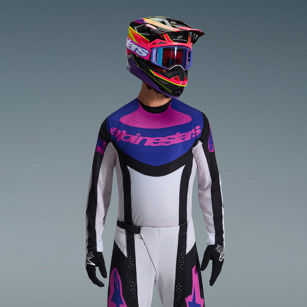Motocross rider wearing Alpinestars 2026 Techstar Knif Gear Set, Jersey with blue and pink chest graphic and white torso, white pants with black panels and pink logos, multi-colored helmet in purple and pink with matching goggles, black gloves, professional off-road racing gear